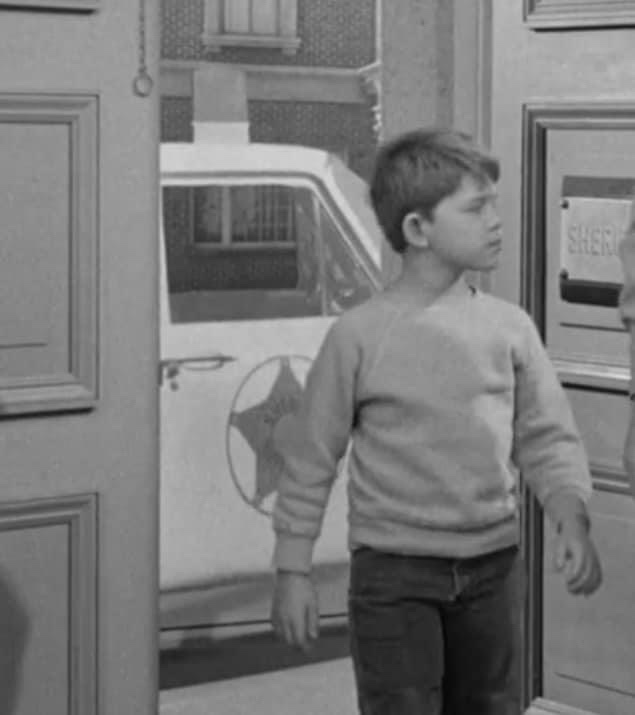 A black-and-white still of a young boy in a sweater standing in a doorway, looking off to the side with a serious…