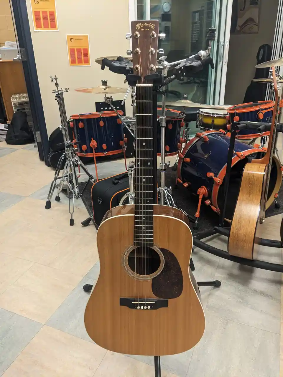 A Martin & Co. acoustic guitar is mounted vertically on a stand in a music room, with drum kits and other instruments…