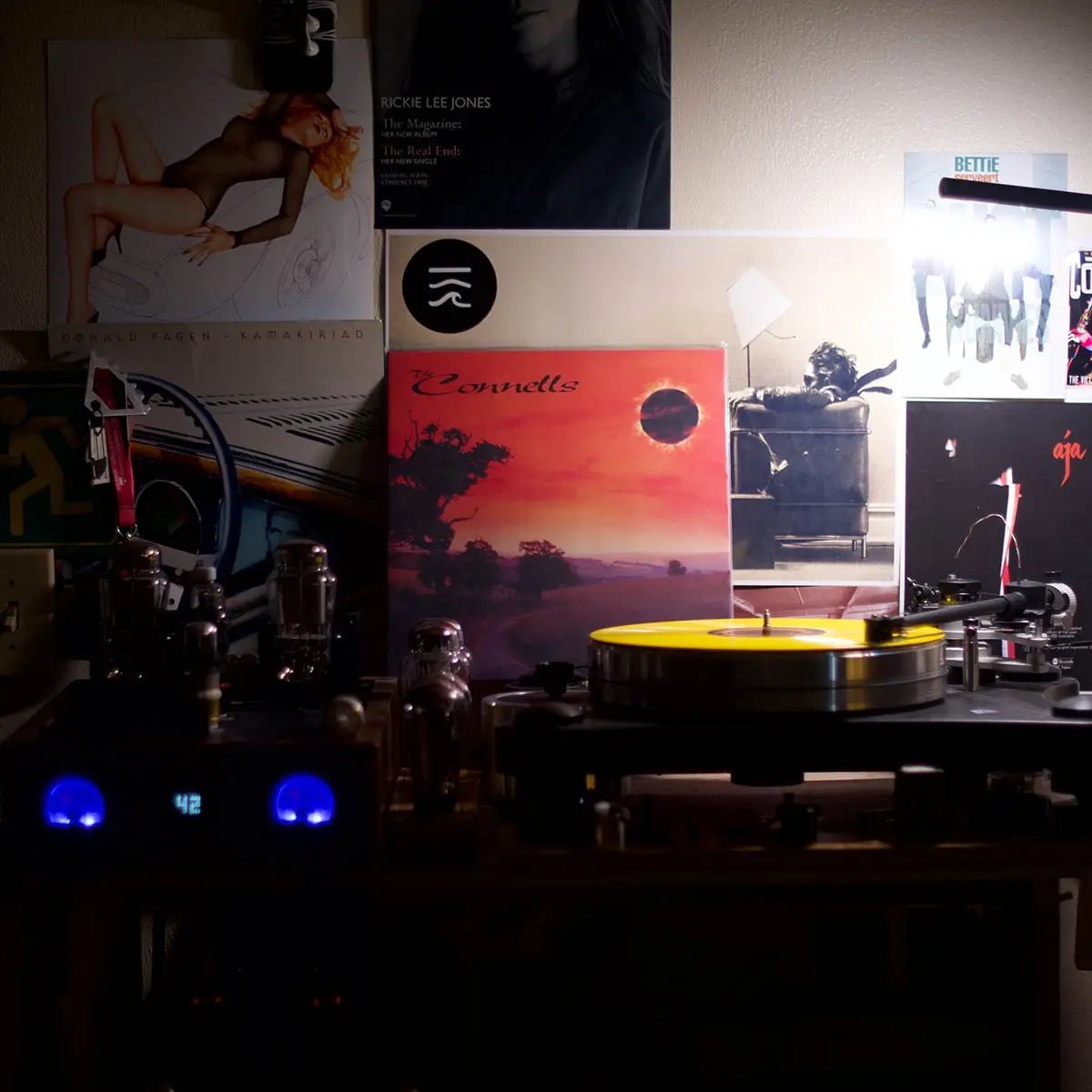 A dimly lit room features a turntable with a yellow vinyl record spinning, surrounded by vintage audio equipment with…