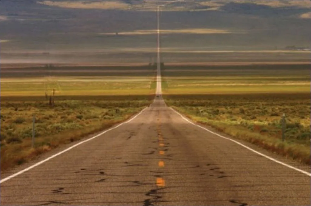 A long, straight, cracked asphalt road stretches into the horizon through a vast, flat, arid landscape under a cloudy sky…