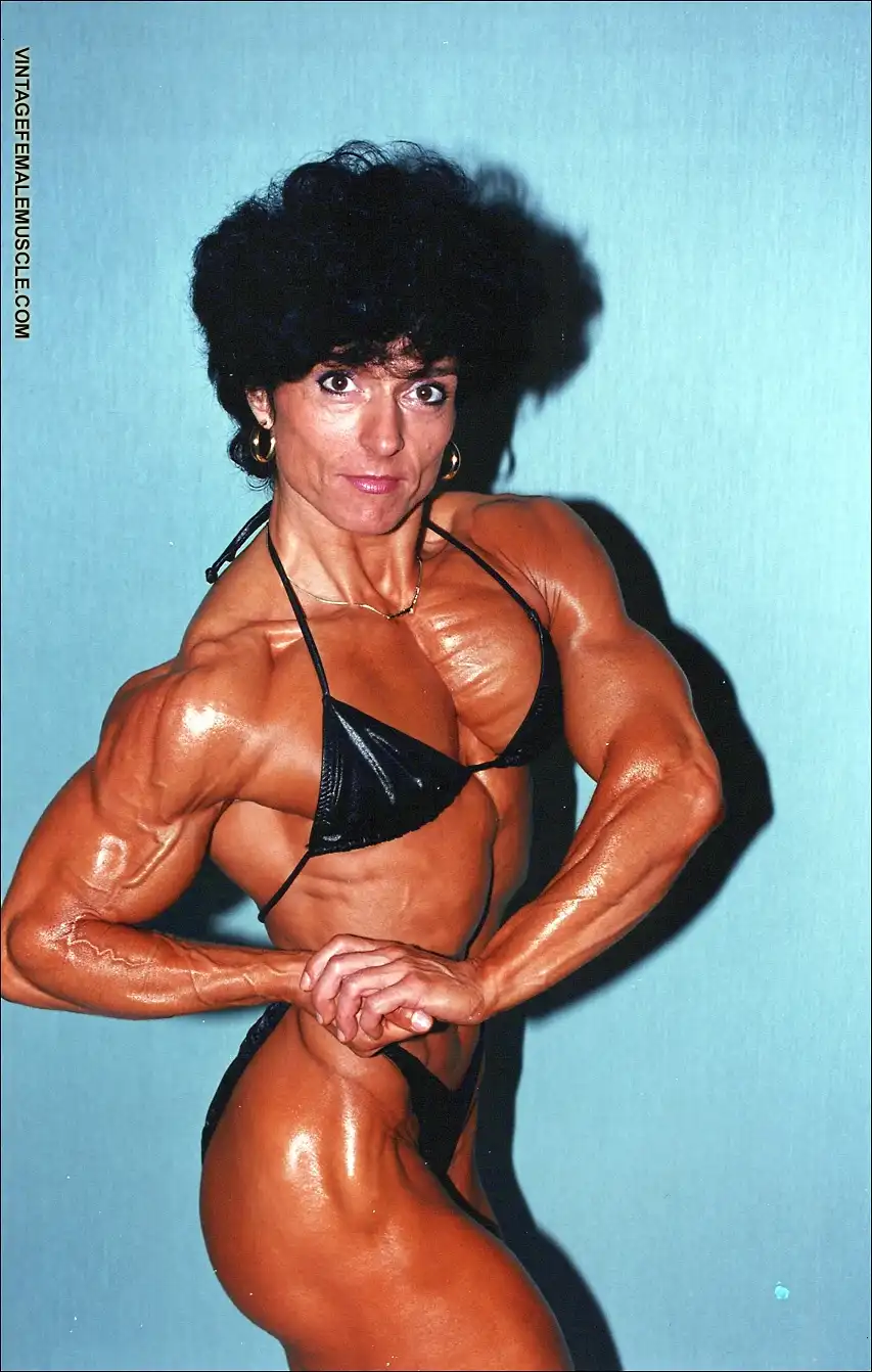 A muscular woman with dark curly hair and gold hoop earrings poses in a black bikini against a blue background, flexing her…