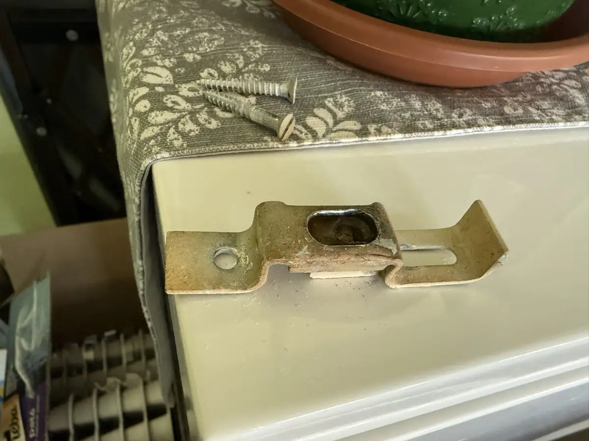 A close-up photo shows a rusty metal latch attached to a cream-colored fridge door, with two screws lying nearby on a…