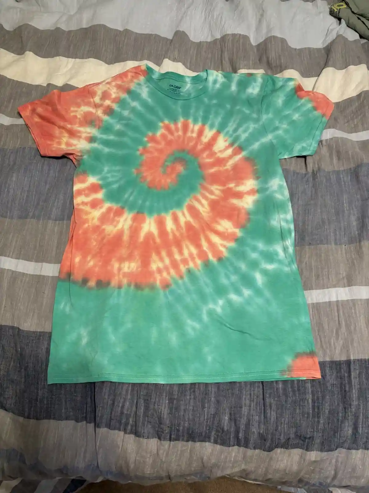 A green and orange tie-dye t-shirt with a spiral pattern lies flat on a striped bedspread. The shirt has a visible 'GILDAN'…