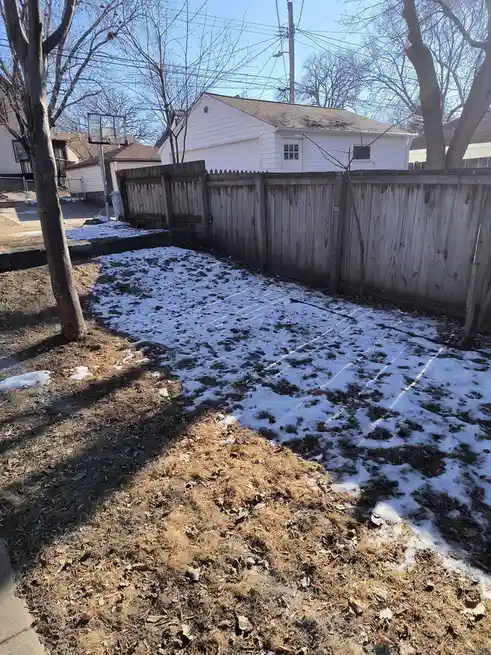 A backyard with patches of melting snow shows faint white lines drawn on the ground, resembling a basketball court, with a…