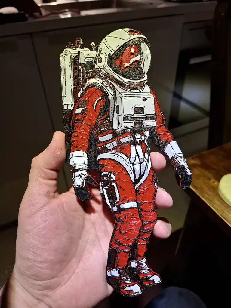 A hand holds a small, detailed figurine of an astronaut in a red and white spacesuit, which appears to be made from…