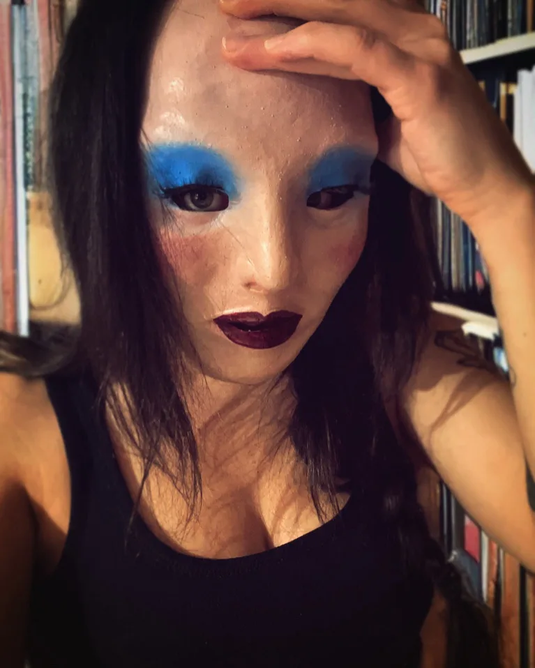 A person with dark hair and a black tank top is shown with a pale, textured mask covering their face, featuring bright blue…