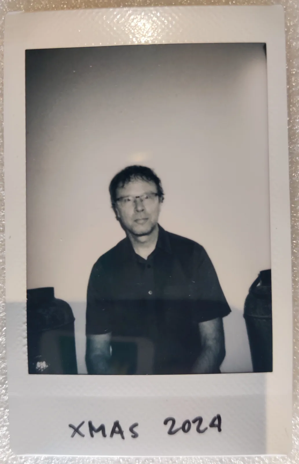 A black-and-white Polaroid photo shows a man with glasses and a dark polo shirt standing between two large, dark containers…