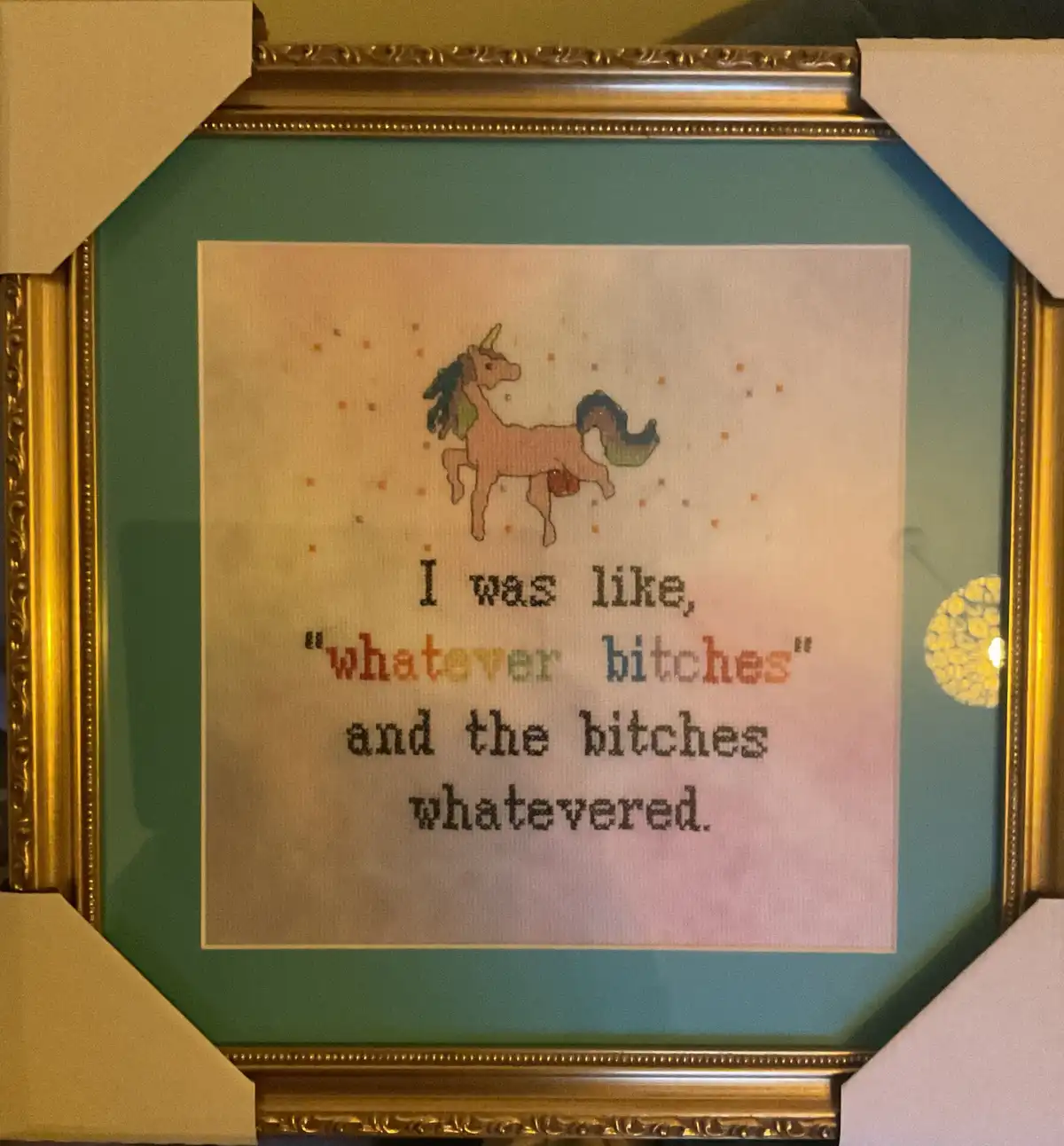 A framed cross-stitch artwork features a cartoon unicorn with a rainbow mane and tail, set against a background of scattered…