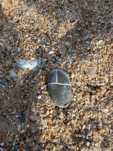 A smooth, dark gray rock lies on a bed of multicolored pebbles and sand, marked with white lines forming a cross and a small…