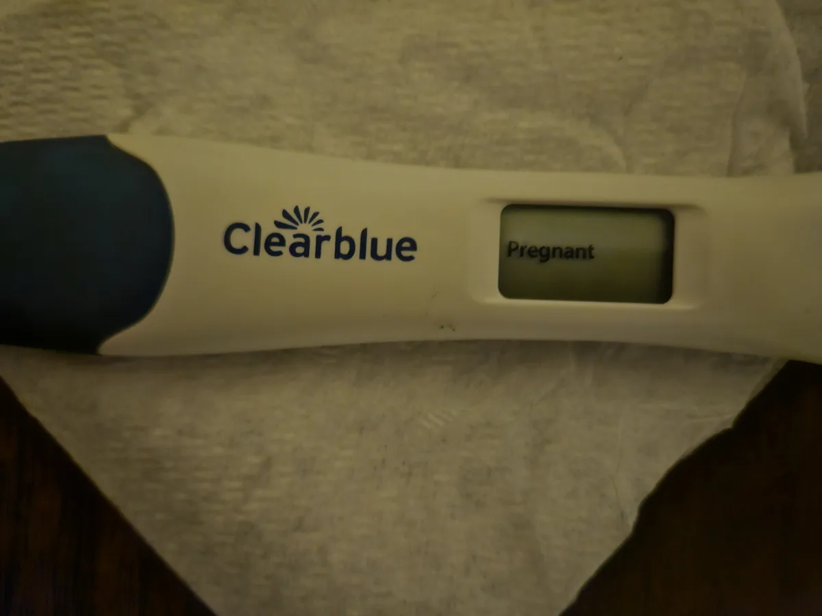A close-up photo shows a Clearblue digital pregnancy test resting on a textured white surface, with its screen clearly…