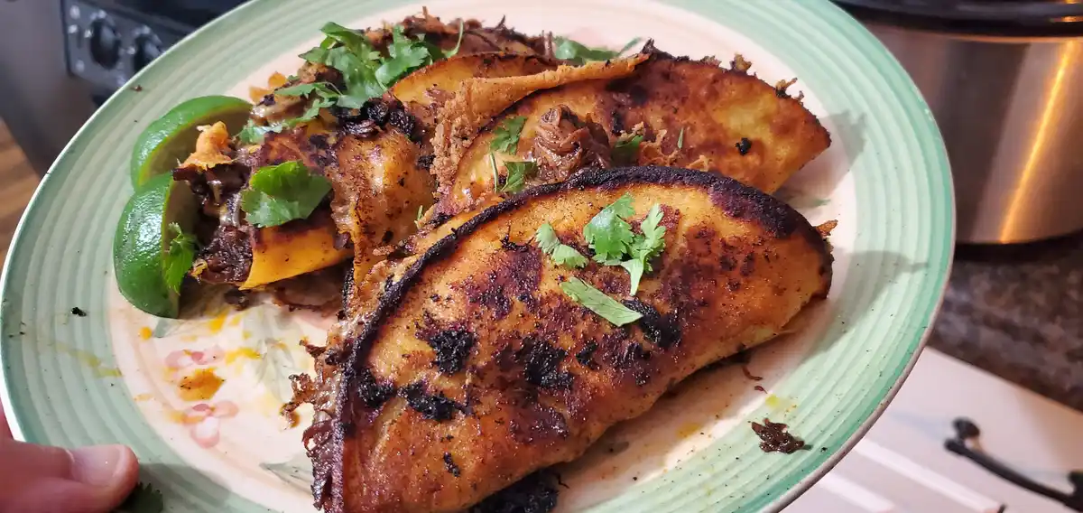 A close-up photo shows a plate of three tacos with heavily charred, blackened tortillas, garnished with cilantro and lime…