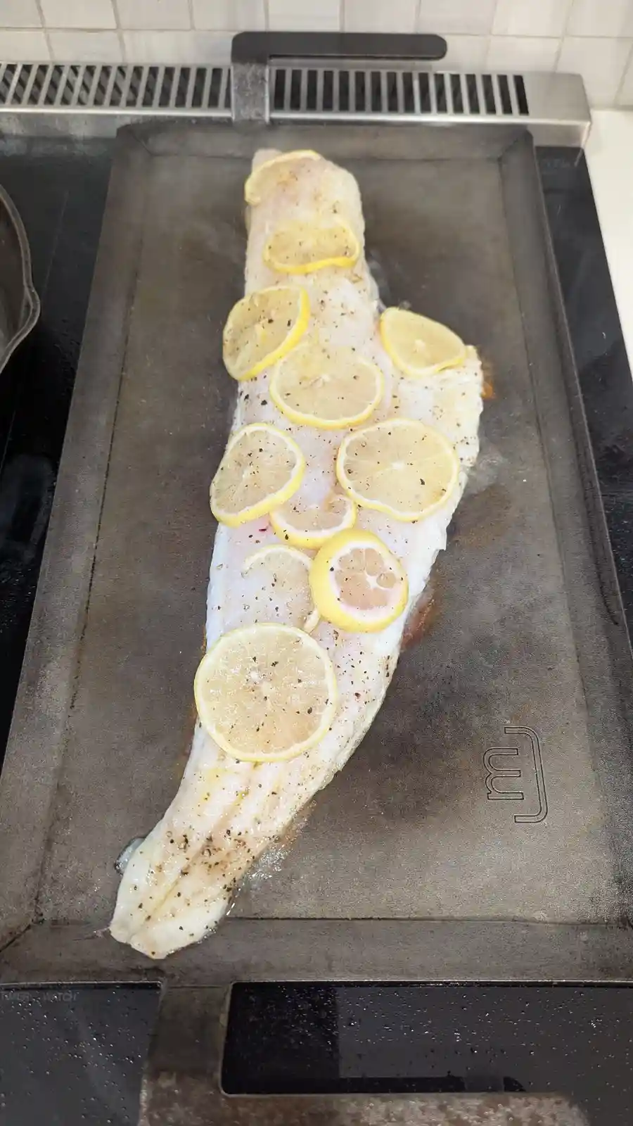 A long, raw fish fillet is cooking on a metal griddle, topped with numerous circular slices of lemon and seasoned with black…