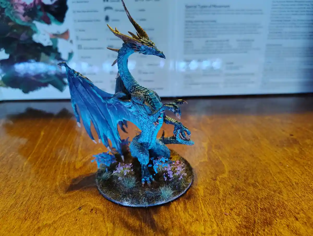 A blue-painted miniature dragon figurine with gold horns and wings stands on a circular base with purple and green terrain…