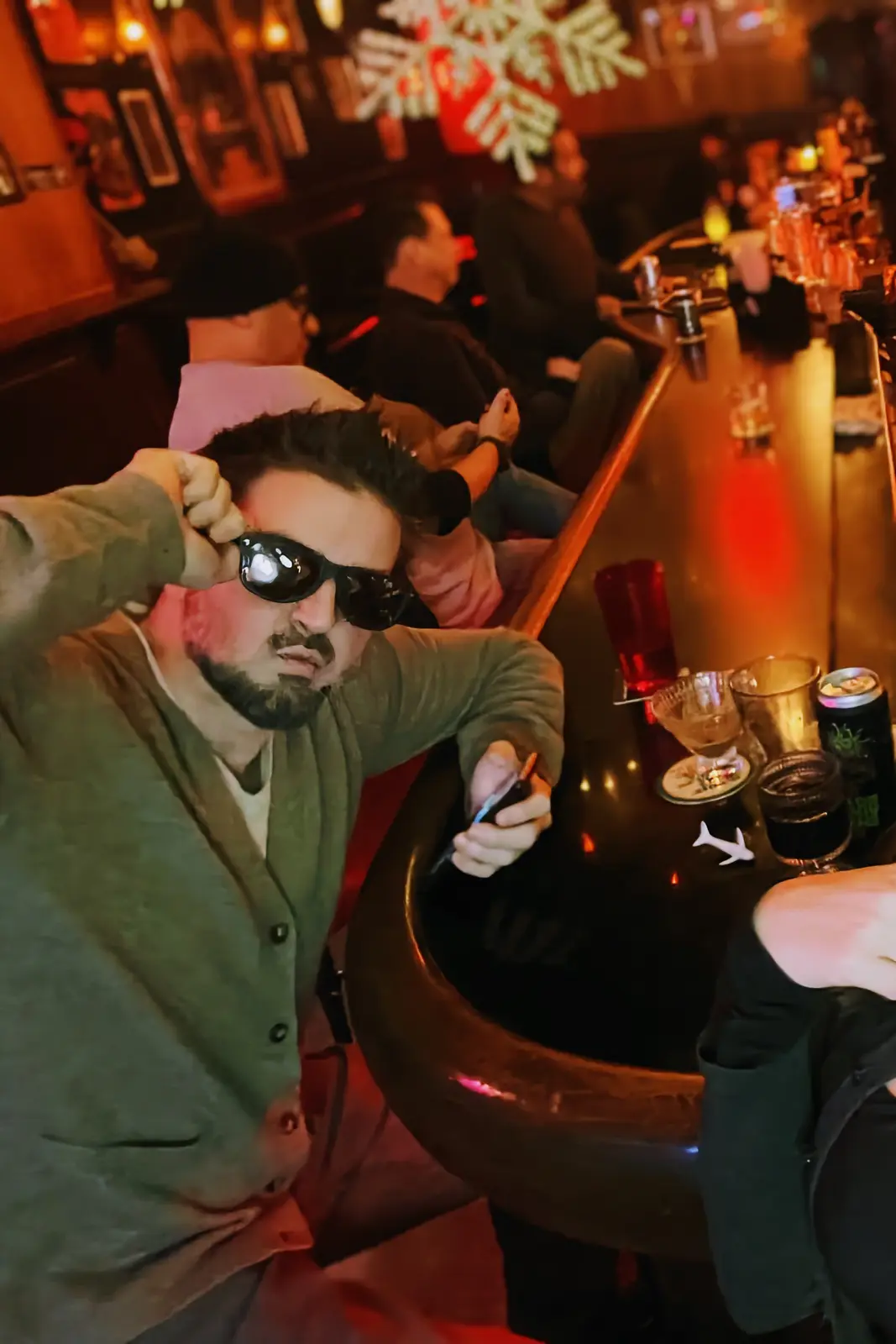 A man with a beard and sunglasses sits at a dimly lit bar, adjusting his glasses while holding a phone. The bar has a warm…
