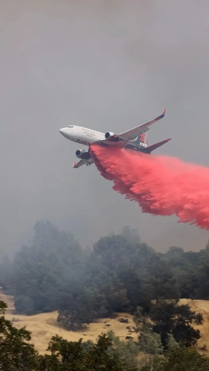 a white coulson air tanker aircraft, marked with 'T-427', flies low over a hazy, smoke-filled landscape with trees and dry…
