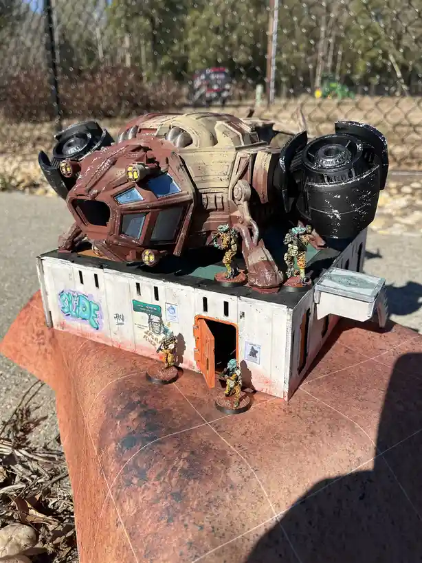 A miniature sci-fi diorama shows a heavily weathered, red and black armored vehicle perched atop a small, white…