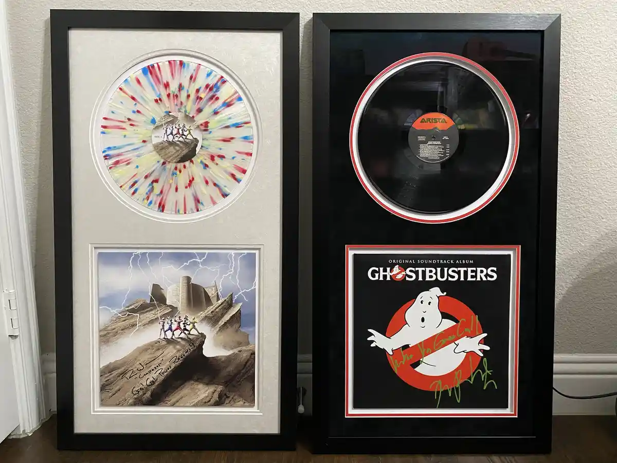 Two framed music memorabilia pieces stand side by side. The left frame contains a colorful, splatter-painted vinyl record…