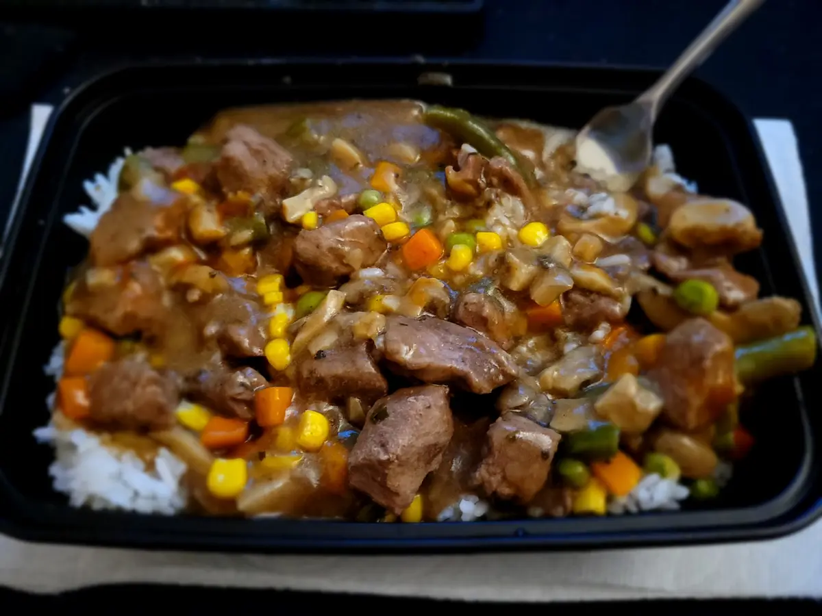 A close-up photo of a black takeout container filled with rice topped with a brown gravy-based stew containing chunks of…