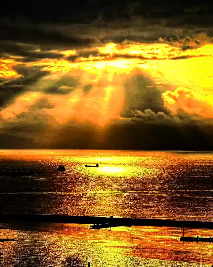 A dramatic sunset over water with golden light breaking through dark clouds, silhouetting boats and a pier. The overlaid…