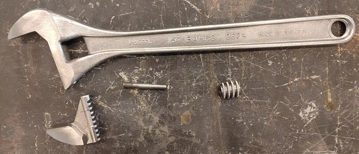 A disassembled adjustable wrench lies on a scratched metal workbench, with its jaw components and a small threaded rod…
