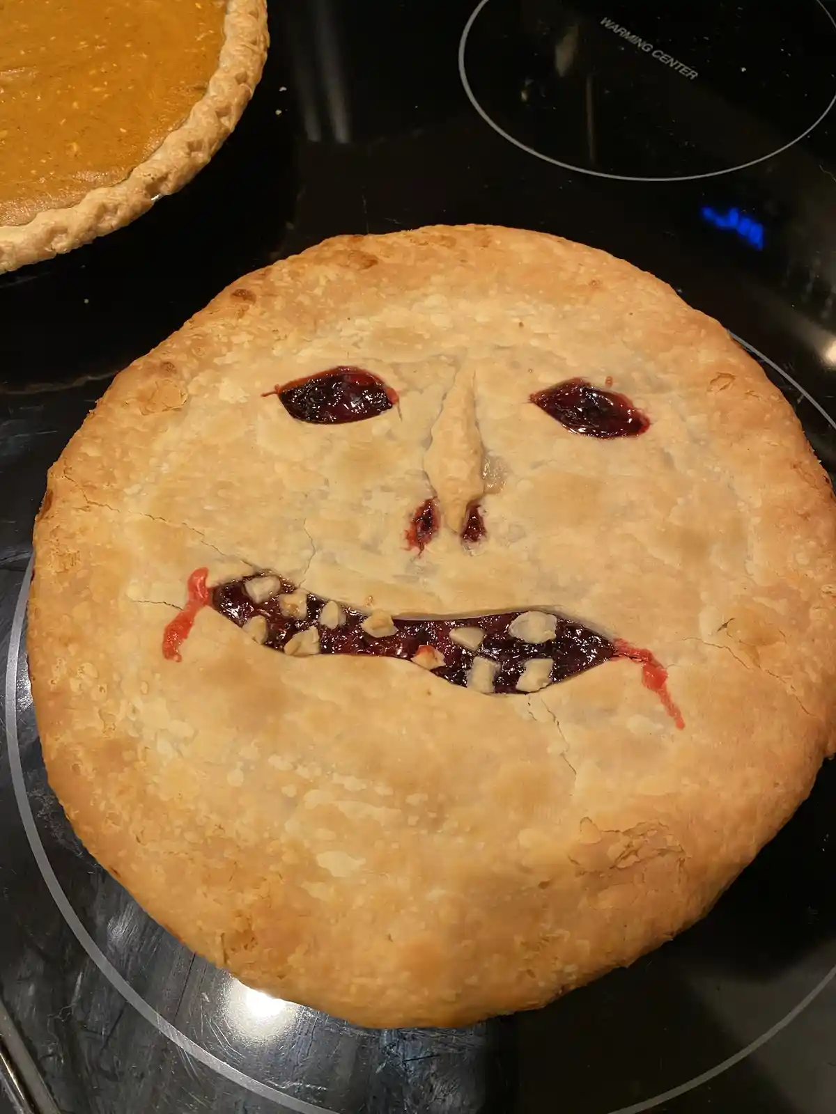 A golden-brown baked pie sits on a black stovetop, its crust decorated with two dark red jam eyes, a small triangular piece…