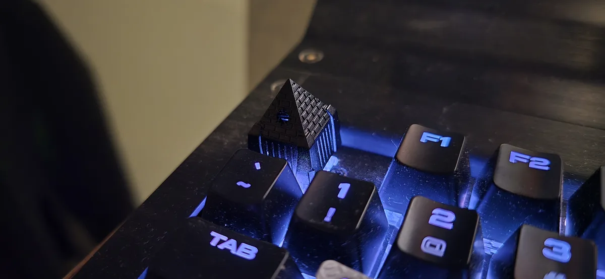 A close-up of a black mechanical keyboard with blue backlighting, where the escape key has been replaced with a detailed…