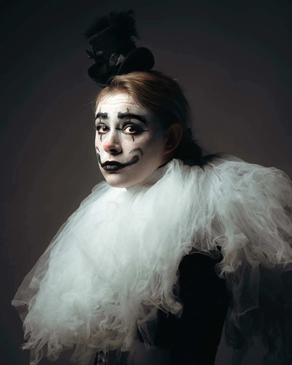 A person with white face paint, black tear-like markings under their eyes, and a black mustache is dressed in a white…