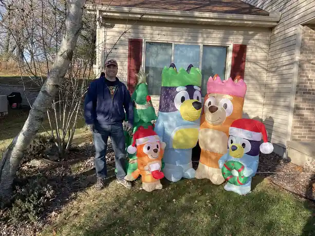 A man stands smiling in a yard next to several large inflatable holiday decorations featuring characters from the children's…