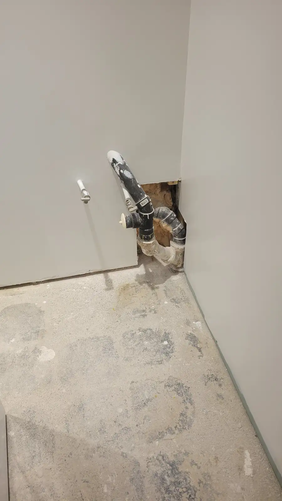 A corner of a room with unfinished drywall walls and a concrete floor covered in dust and debris. A black plumbing pipe…