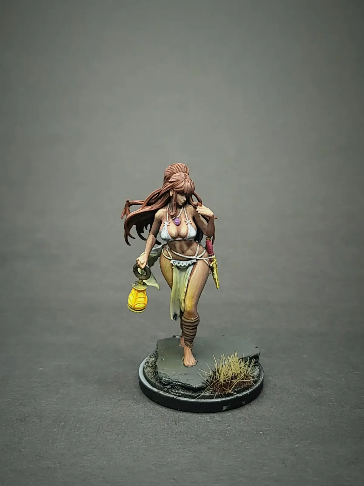 A painted miniature figurine of a woman with long brown hair, wearing a bikini top and skirt, holding a lantern, standing on…