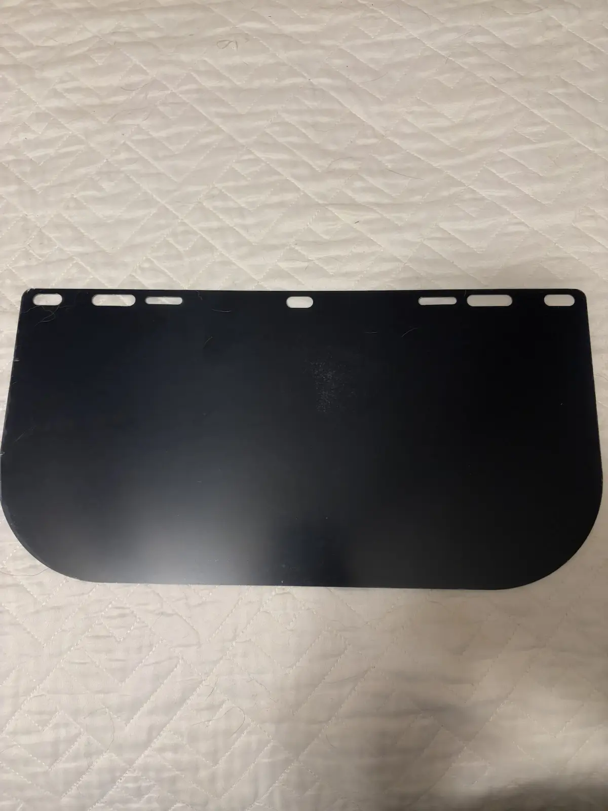 A black, curved face shield with multiple slots along the top edge lies flat on a white quilted surface. The overlaid text…