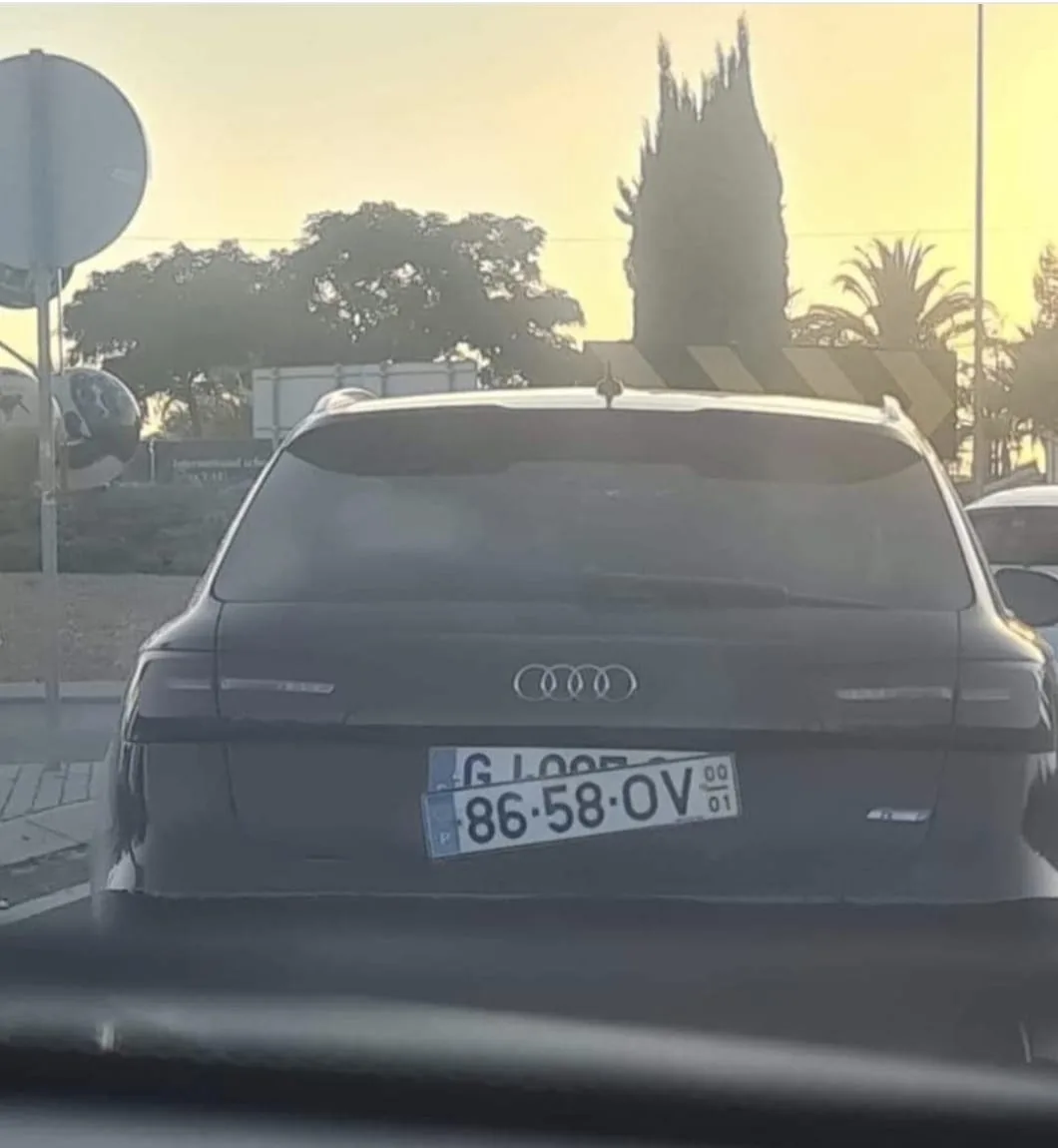 A rear view of a dark Audi car with a license plate that reads 'G LOOB 86-58-OV' under a golden sunset sky. The car is…