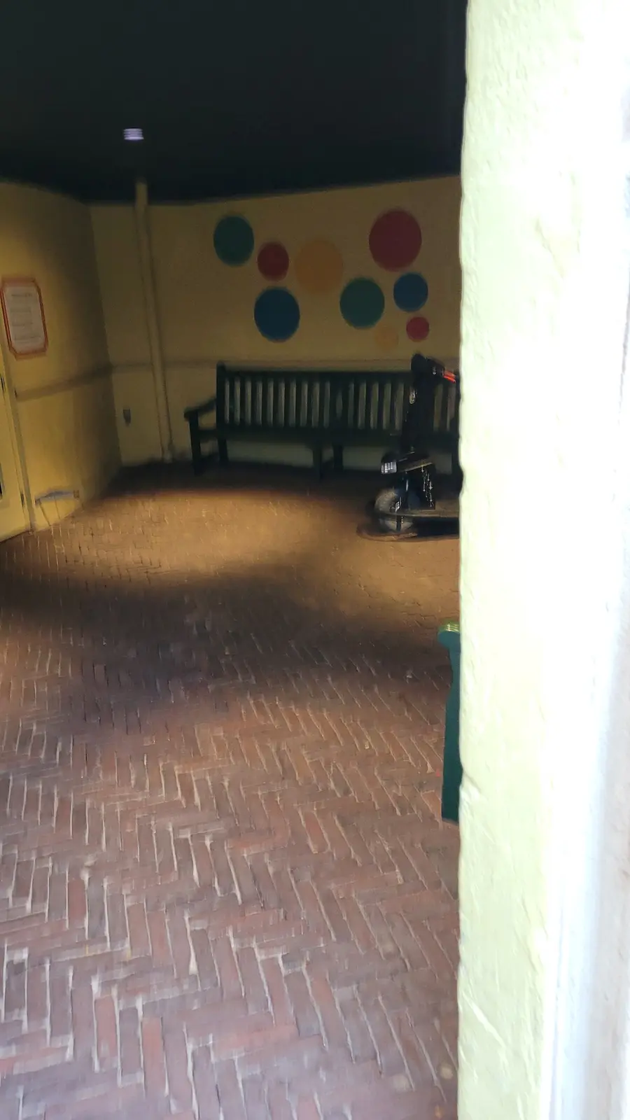 A dimly lit, empty hallway with a brick floor, a bench, and colorful circular wall decorations. A mobility scooter is parked…