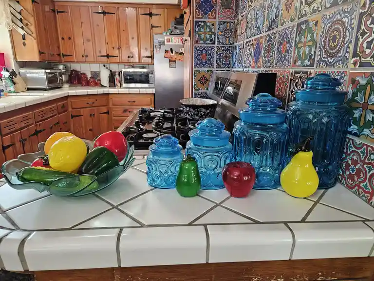 A kitchen counter displays several large blue glass canisters and smaller colorful glass fruit-shaped ornaments, including a…
