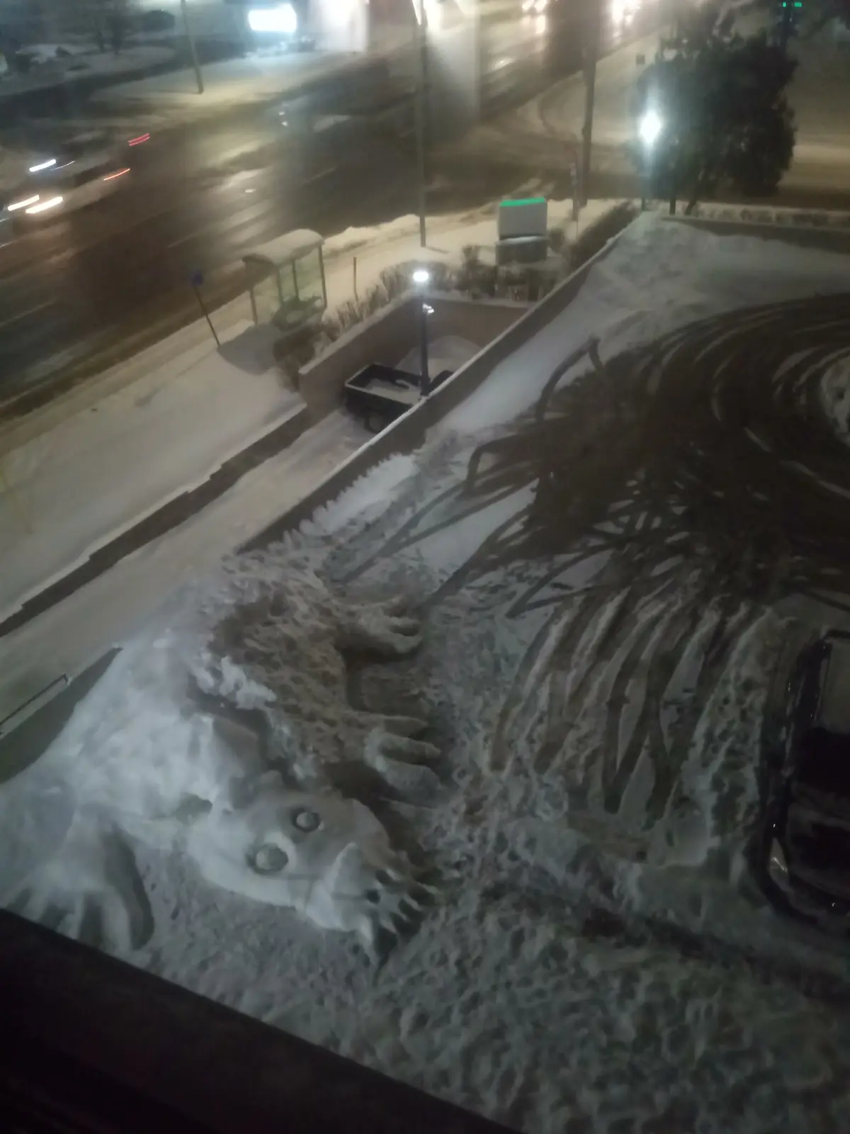 A nighttime photo taken from a high vantage point shows a large, snow-covered creature with a face, claws, and skeletal-like…
