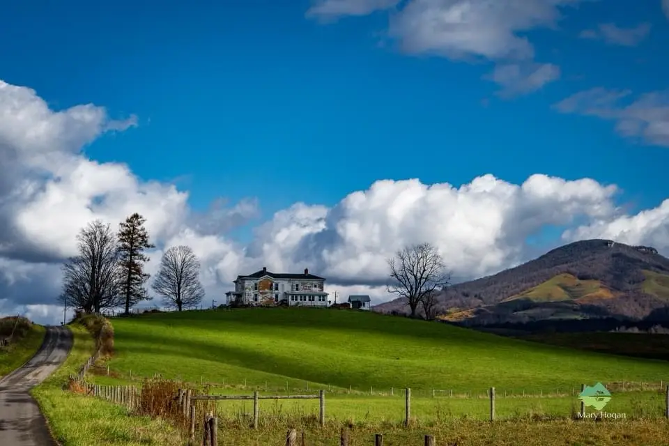 A picturesque rural scene shows a white two-story house perched atop a lush green hill under a bright blue sky with fluffy…