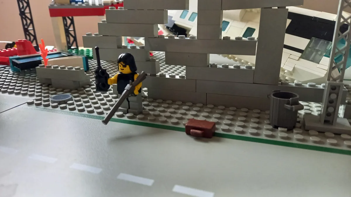 A lego minifigure with a black helmet and vest holds a rifle and a walkie-talkie, standing beside a red briefcase on a grey…