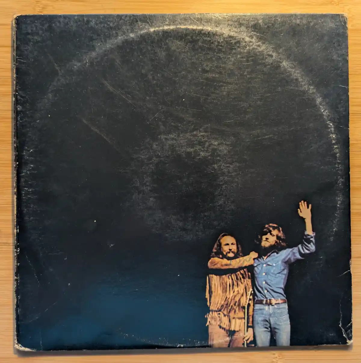 A heavily scratched and worn black vinyl record sleeve shows two men, one in a fringed jacket and the other in a blue shirt…
