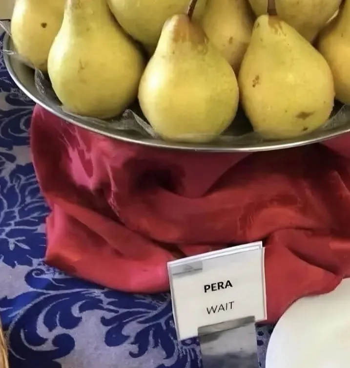 A metal bowl filled with yellow pears sits on a red cloth atop a blue patterned tablecloth. A small white sign in front…