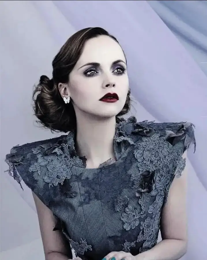 A woman with dark, dramatic eye makeup and red lipstick stares intently upward, wearing a dark, ornate, lace-adorned dress…