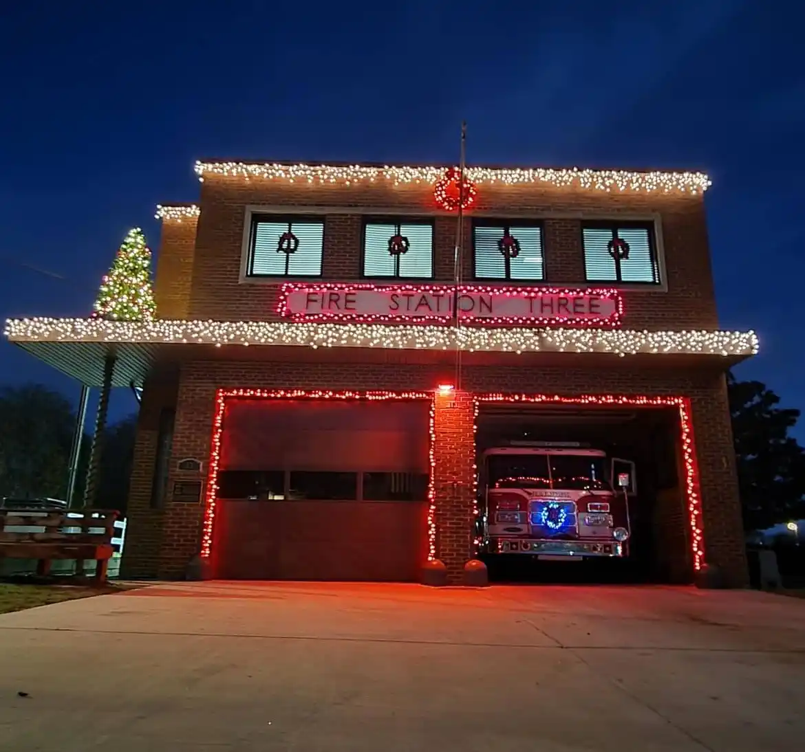 A brick fire station at night is heavily decorated with white and red Christmas lights outlining the roofline, windows, and…