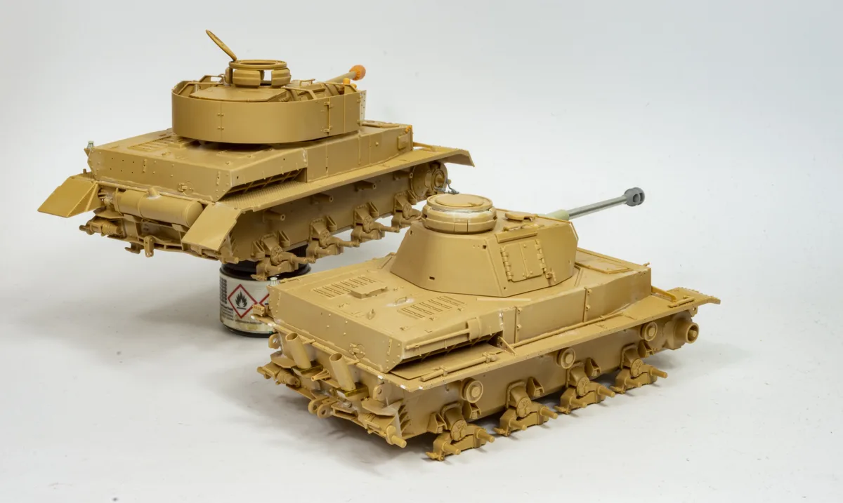 Two unpainted tan plastic model tanks sit on a white surface, one slightly behind the other. The rear tank rests on a small…