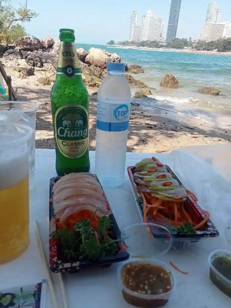 A table on a beach with a bottle of Chang beer, a bottle of Top Aquafina water, and two plastic containers of Thai seafood…