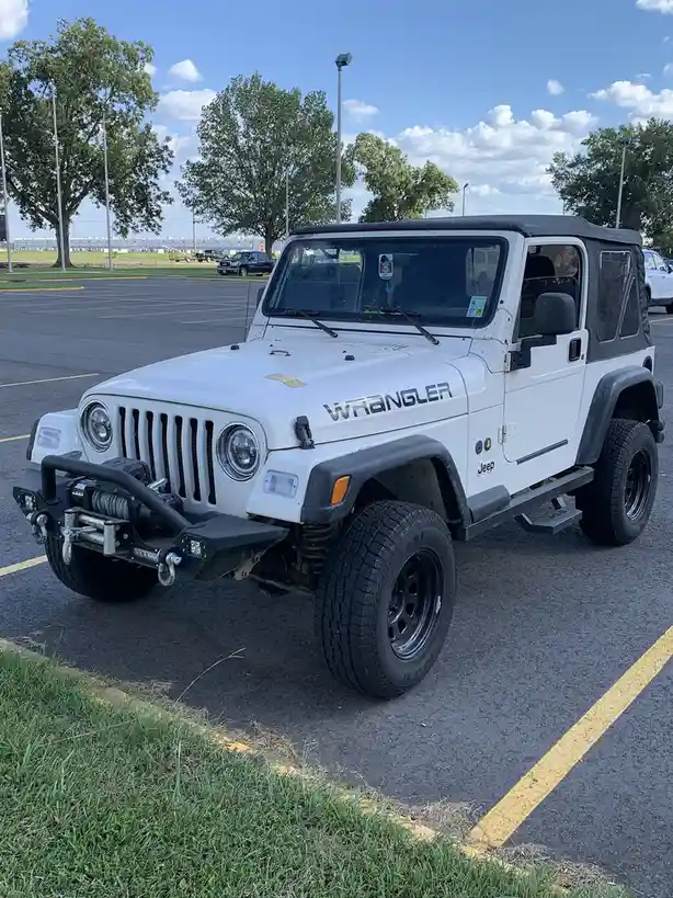 A white Jeep Wrangler with black trim and a soft top is parked in an asphalt lot under a partly cloudy sky. The vehicle…