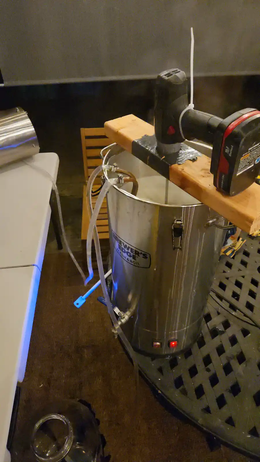 A silver 'Brewer's Edge' brewing kettle sits on a metal grate, with a power drill mounted on a wooden plank above it…