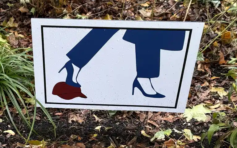A white rectangular sign with a black border, covered in water droplets, is stuck in dirt and leaves. It shows a blue…