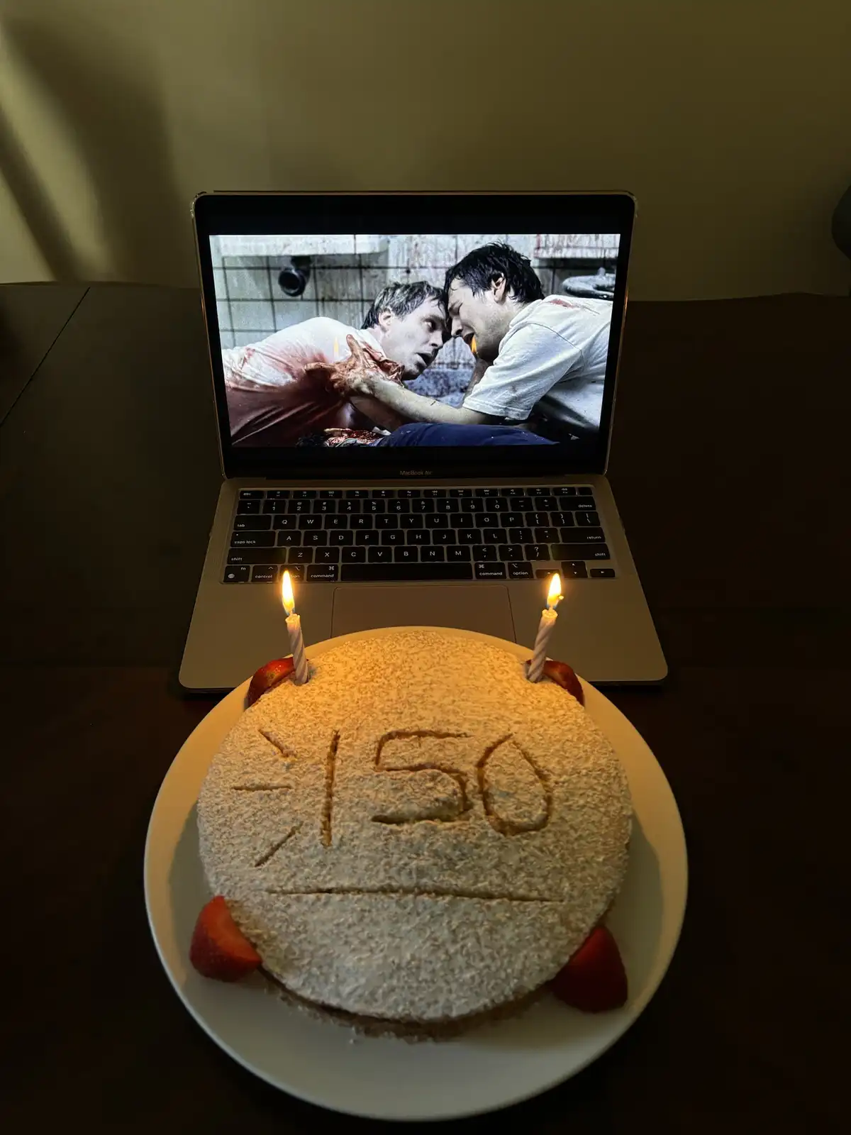A birthday cake with two lit candles and the number '150' written in icing sits in front of a laptop displaying a graphic…