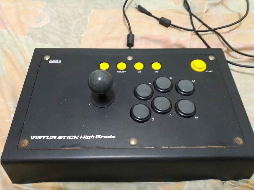 A black Sega Virtua Stick High Grade arcade fighting game controller with a central joystick, eight black buttons labeled…
