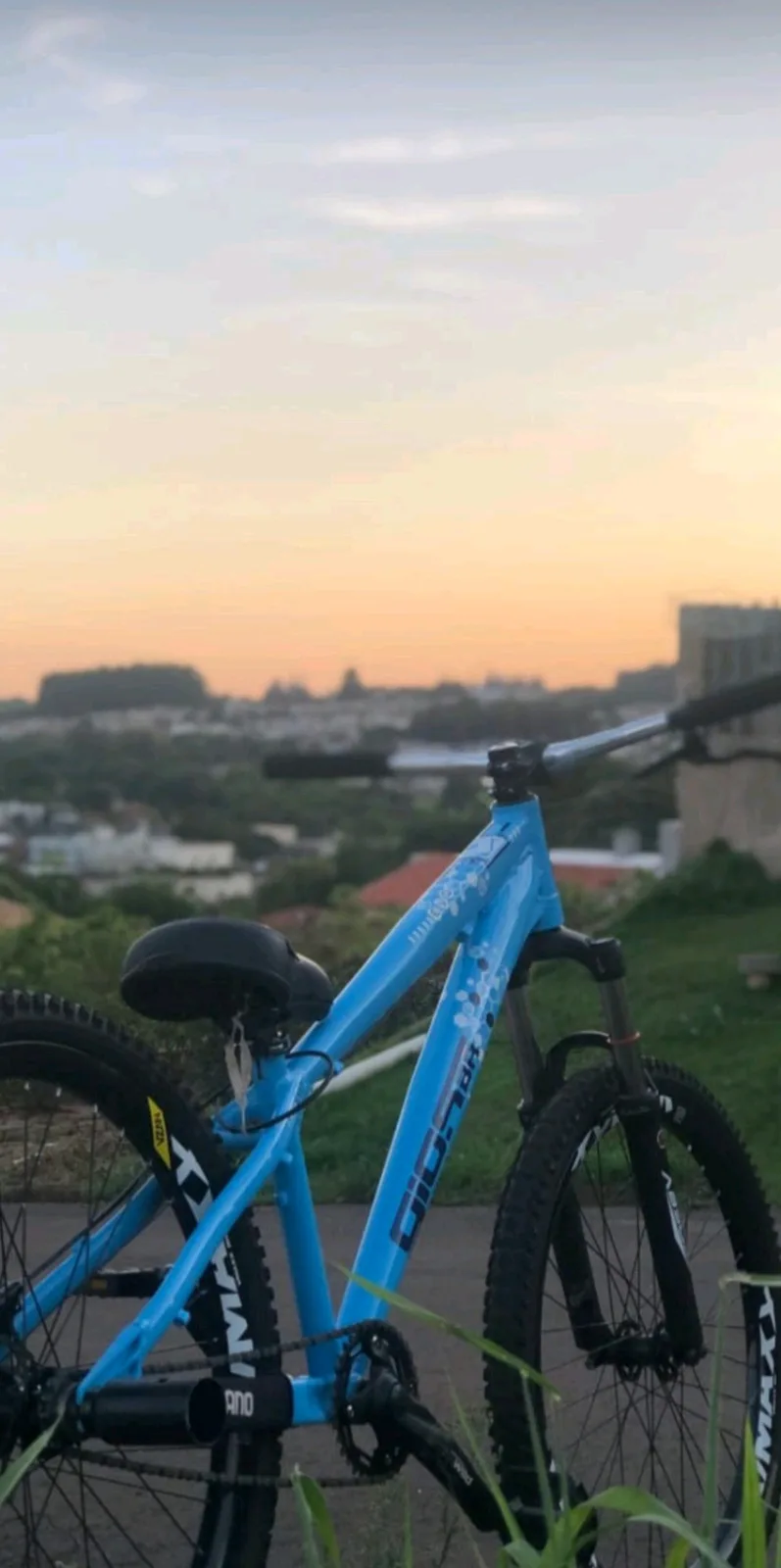 A bright blue mountain bike is parked on a paved path with a scenic sunset over a cityscape in the background. The bike has…