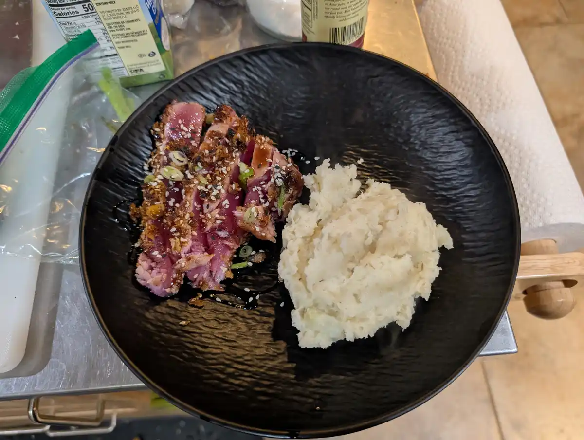 A black textured plate holds sliced seared tuna with sesame seeds and green onions next to a scoop of mashed potatoes. The…